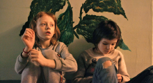 Two children sitting in front of a yellow wall and a large green plant