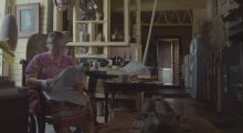 An older woman sitting on a chair, reading a newspaper, in a living room.
