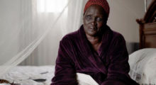 A middle-aged Black woman in a mauve bathrobe and red headscarf on a white bedspread