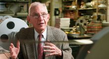 An old white man with glasses and a light brown suit in front of film reels at a film laboratory