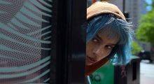 A Black woman with blue hair and an tan hat peering around the corner of a building