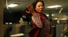 an Asian woman with long black hair standing, arm outstretched, amidst a swirl of papers in an office