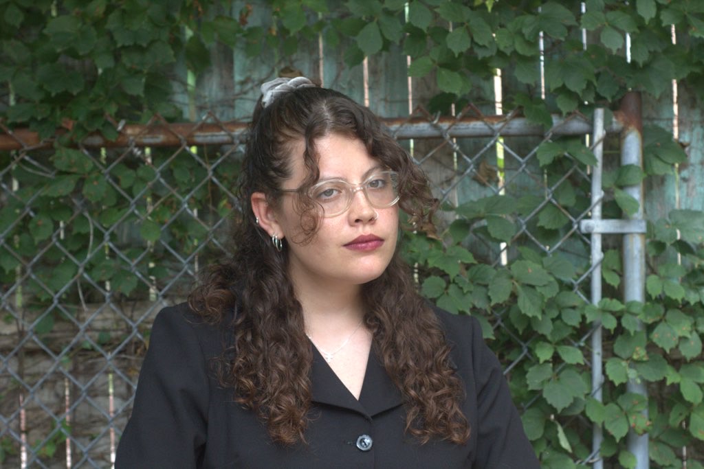 A young woman with brown hair and a black blouse in front of a wall of plants