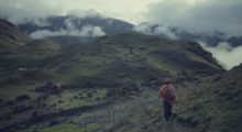 A wide shot of a man walking in a mountainous plain with clouds overhead