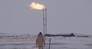 A figure in a heavy winter coat stands in front of an oil pipeline shrouded in fresh snowfall.