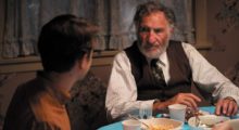 The back of a young boy's head with brown hair is turned toward an elderly man wearing a white shirt with pinstripes and a brown sweater vest. They are sitting at a dining table during dinnertime.