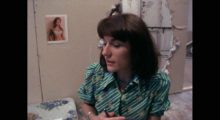 A middle-aged woman with brown hair and a green top in a room with beige walls