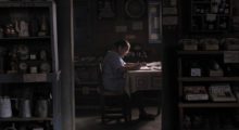A woman in a blue floral house dress sits at a wooden dining table with a pen and paper, surrounded by curated clutter in wood-paneled room.