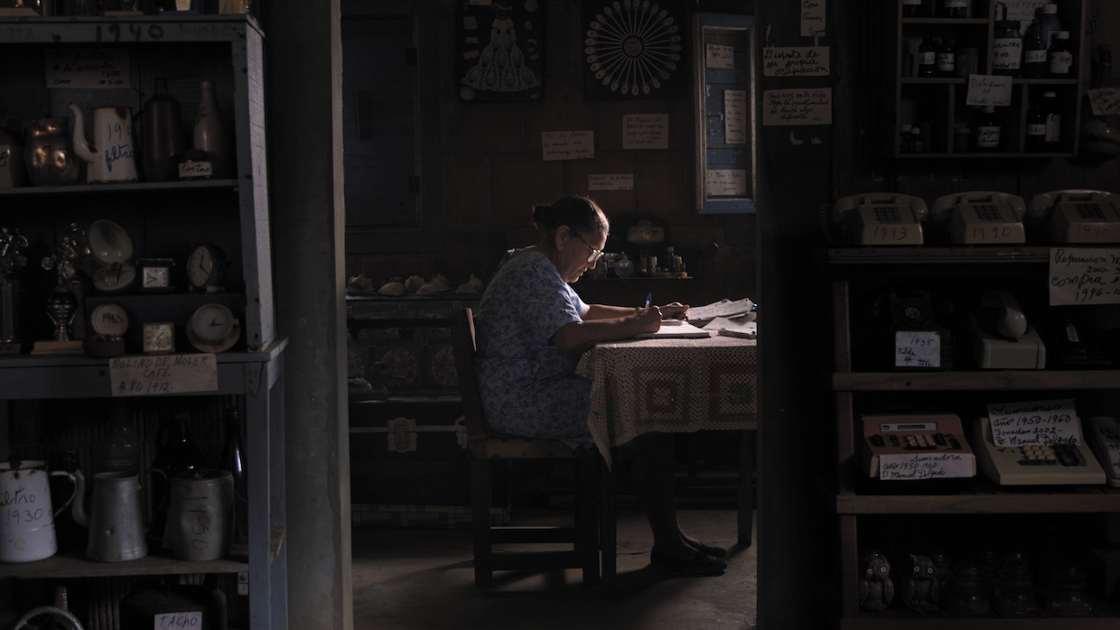 A woman in a blue floral house dress sits at a wooden dining table with a pen and paper, surrounded by curated clutter in wood-paneled room.
