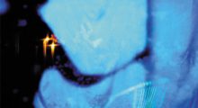 An abstract, blue-tinted figure is superimposed over a chair and positioned next to faint candlelight.