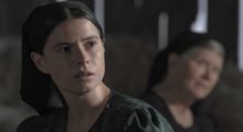 A young, white Mennonite woman looks in the distance, with an older woman in the background behind her.