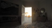 Inside of a house filled with sand, the front door is wide open, revealing the shadow of a man standing far off in the background.