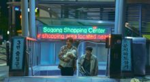 Two Korean men ascend an escalator in a shopping center, with the older of the two men holding a baby in his arms.