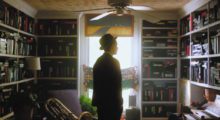 A man wearing a suit and top hat stands in front of walls lined with built-in bookcases, each shelf filled with VHS tapes.