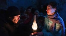 A man and a woman sit opposite each other wearing ratty but warm clothes. An oil lamp burns between them.