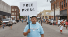 A young man wearing a blue t-shirt holds a sign that reads "free press."