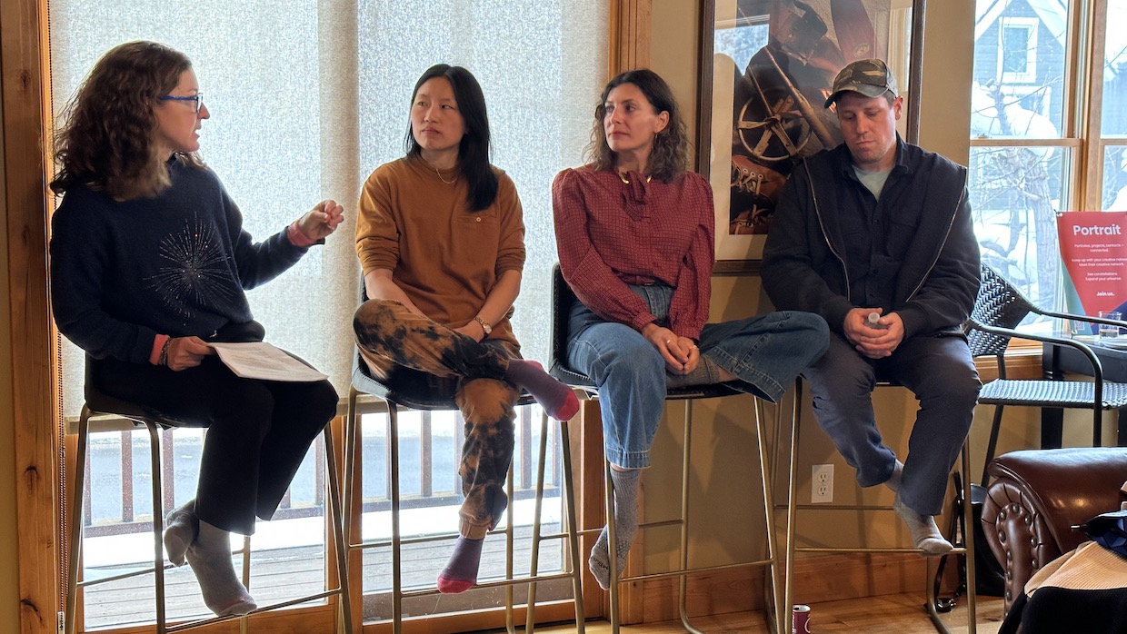 Three women and one man sit on tall stools in a wood-paneled room with large windows.