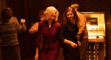 Two women, one blonde and one brunette, wearing 1960s-era outfits dance in front of a jukebox.