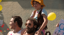 A young girl wearing a brown vest and bucket hat sits on her bearded father's shoulders as a parade passes them by.