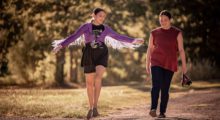 Two young women walk along a dirt path in a park. One wears a purple fringe jacket, a band t-shirt and black bike shorts with checkered vans. The other wears a sleeveless red t-shirt, black jeans and sandals.