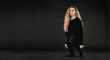 A woman with long, curly blond hair wears all black and stands amid a dark gray background. She has a rare condition where her legs are disproportionally short compared to her body.