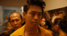 A young man stands as people gather around him, an elderly man can be seen in the background directly behind him.