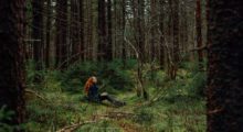 A woman with long, wavy red hair sits in a forrest surrounded by tall trees and lush grass.