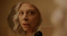 An older woman with gray hair looks at her reflection in a compact mirror.