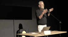 A man with white hair and a trimmed beard stands in front of a desk giving a lecture.