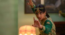 A young woman wears a green gown embroildered with gold details. She wears an ornate gold headpiece and holds her hands up in combat.