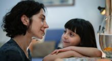 A woman with cropped, curly brown hair sits next to a young girl with straight black hair and bangs. They gaze at each other.