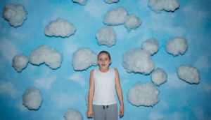 A young girl with blond hair wears a white tank top and gray sweatpants and stands in front of a wall with a blue sky and clouds painted on it.