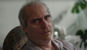 A white man with gray disheveled hair wears a gray button-up pajama top and looks off into the distance.