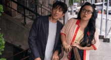 An Asian man and woman stand on the street, leaning slightly to their left while staring at something in the distance.