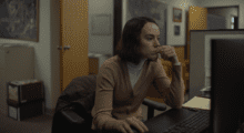 A woman with a shoulder-length bob haircut wears a camel-colored cardigan and sits in front of a computer at a desk in an office. She touches her fingers to her lips absentmindedly.