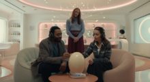 A young couple sits in a retro-futuristic room with pink-hued underlights. An egg-shaped object, perhaps really an egg, sits on a small wooden table in front of them. Behind them, a woman with shoulder-length blond hair looks over their shoulders.