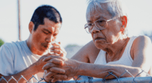 A young man wearing a gray t-shirt smokes a cigarette while an elderly man in a white tank top leans on a chainlink fence behind him.