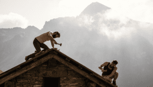 Two men work on a house's roof with hammers with the mountains of the Swiss Alps in the background.