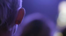 Someone's right ear featuring a single black horseshoe-shaped hoop. They are bathed in indigo-colored lighting, likely at a music venue.