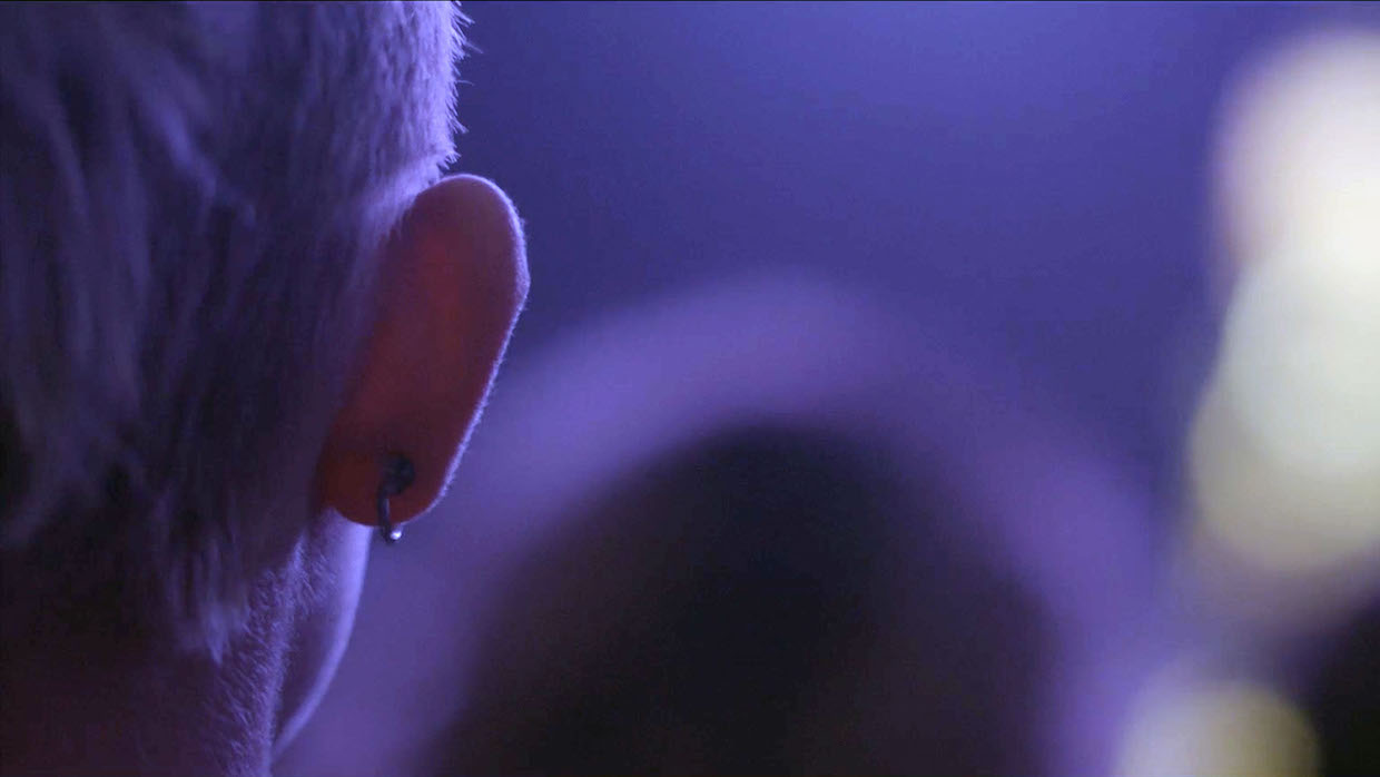 Someone's right ear featuring a single black horseshoe-shaped hoop. They are bathed in indigo-colored lighting, likely at a music venue.