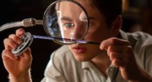 A boy wearing a tan polo shirt examines a strip of film under a magnifying glass.