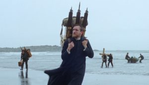A man dressed in priest's robes walks along the sandy shores of the Icelandic coast, with a wooden chest strapped to his back. Several men drag a rowboat onto the shore in the background.
