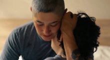 A person wearing a gray t-shirt with a shaved head holds their child with brown curly hair close to their body.