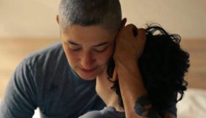 A person wearing a gray t-shirt with a shaved head holds their child with brown curly hair close to their body.