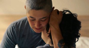 A person wearing a gray t-shirt with a shaved head holds their child with brown curly hair close to their body.