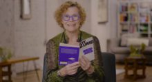 A woman with short, curly red hair sits in a chair with an open copy of Are You There, God? It's Me, Margaret.