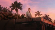 A man in a white linen suit stands on a roof with a bright pink sky and palm trees in the background.