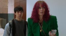 A young man in a gray t-shirt and light gray zip-up hoodie stands next to a taller woman with shoulder-length, curly red hair wearing a green suit.