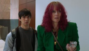 A young man in a gray t-shirt and light gray zip-up hoodie stands next to a taller woman with shoulder-length, curly red hair wearing a green suit.