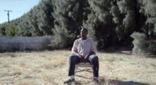 A Black man sits in a chair outdoors, with dead grass covering the ground and an imposing line of thick trees cutting through the background.